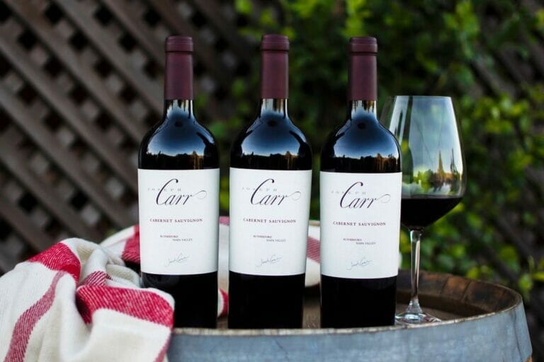 three bottles of cabernet sauvignon, wine glass and towel sitting on barrel outside