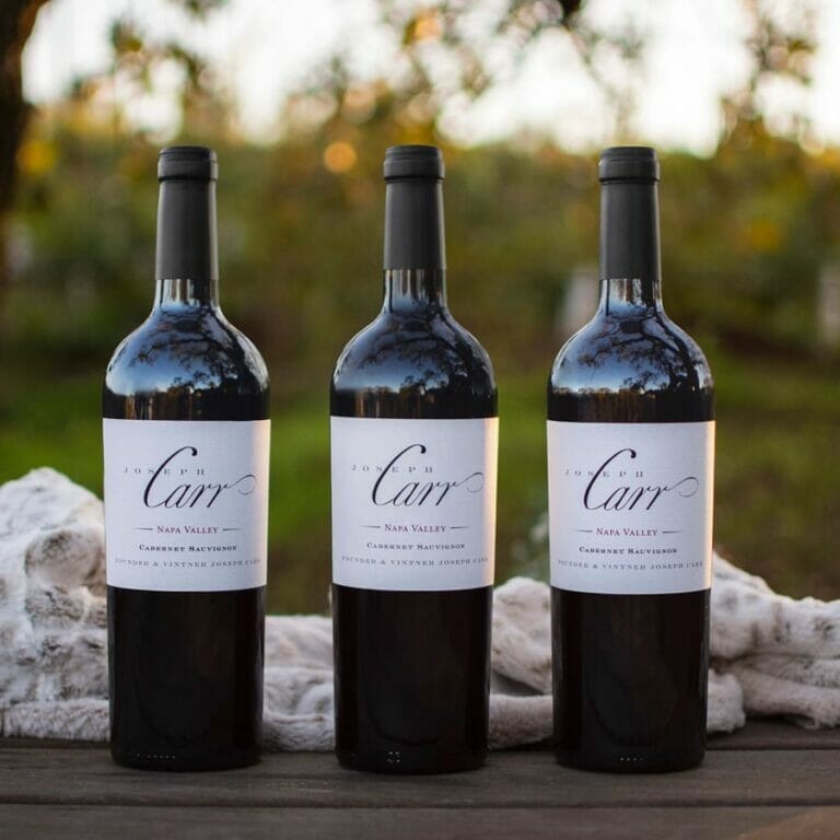 three bottles of carr cabernet sauvignon sitting on wooden table in front of blanket and trees